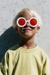 © Andrii Zastrozhnov - Blond child wearing round white sunglasses with red lenses against concrete wall for summer poster and kids fashion banner