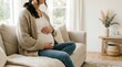 © asih - Pregnant woman sitting on a beige couch in a bright room cradling her belly with hands