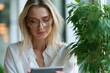 © PixelPulse Creative - Portrait of a young blonde woman wearing glasses and a white shirt, looking down at a tablet near a green plant