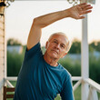 © SYATRI RAWU - Active Senior Caucasian Man Exercising Outdoors on a Sunny Porch During Sunset Performing Side Stretch Exercises for Healthy Aging and Wellness.