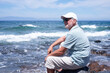 © luciano - Senior man relaxing on a rocky beach by the ocean, wearing a cap and sunglasses, enjoying the sunny day and contemplating the vast sea