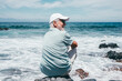 © luciano - Senior man relaxing on a rocky beach, enjoying ocean waves and sun, looking away while thinking about retirement and travel, feeling freedom and happiness