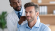 © MargoArt - Smiling handsome man looking forward while professional african american doctor in white coat touches his neck against blurred clinic room background