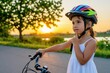 © Vadym - Small girl wears colorful safety helmet riding bike on summer road at sunset. Child touches chin looking thoughtful during outdoor activity near green trees. Cute kid enjoys evening cycle ride in
