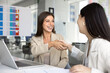 © fizkes - Two professional women smiling and shaking hands seated at desk with laptop and papers in modern office, gesture of mutual respect, successful negotiation outcome, and perspective collaboration. Deal