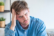 © simona - Young man holding his head in pain sitting on a couch, stressed and suffering from a headache or migraine at home, feeling tired and anxious