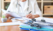 © Nanci - Healthcare professional wearing a stethoscope is reviewing patient records and administration documents, with a stack of paperwork on the desk showing the volume of work