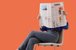 © Pixel-Shot - Woman in blue shirt and jeans reading a fashion newspaper while sitting on a chair, isolated on an orange background