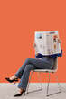 © Pixel-Shot - Woman in blue shirt and jeans reading a fashion magazine while sitting on a chair against an orange background, the persons face is obscured by the open magazine