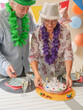 © luciano - Senior adults celebrating a 75th birthday party at home, placing a birthday cake with candles on a decorated table while wearing party hats and leis