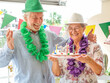© luciano - Happy senior couple celebrating 75th birthday, smiling and holding a birthday cake with candles, wearing party hats and leis, enjoying family celebration indoors