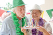 © luciano - Senior couple celebrating 75th birthday, man blowing out candles on cake while smiling wife holds plate, enjoying a happy party moment at home