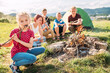 © Soloviova Liudmyla - Portrait of happy little girl in foreground eating roasted marshmallow from stick by warm campfire with friends and green tent visible behind her in sunny mountain meadow during summer camping
