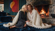 © f_bossa - Couple sits together on a velvet rug before an active stone fireplace. A thick chunky knit blanket covers them as warm fire glow illuminates their faces against dark interior decor.