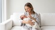 © puja - Smiling mother cradling her swaddled newborn baby on a white sofa in a bright room, ideal for healthcare, motherhood, and family wellness advertisements.
