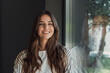 © Daniel - Young woman with long brown hair smiling at the camera, standing indoors next to a large window reflecting trees and nature, wearing a striped shirt