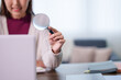 © Audtakorn - Young Asian woman holding magnifying glass while researching with laptop in bright home office with copy space