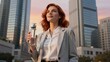 © mihrzn - A confident red-haired woman in a grey blazer holds a water bottle, gazing upward against a backdrop of towering skyscrapers during a warm sunset.