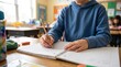 © startang141 - A young student in a blue hoodie diligently writes in a spiral notebook with a pencil at a wooden desk in a bright classroom.