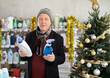 © JackF - Elderly man in winter clothes chooses sprays and detergent in a supermarket on New Year's Eve. Buyer in a household chemicals store against the background of a Christmas tree