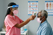 © Pete - African American female nurse with face shield, mask uses infrared thermometer to check body heat of senior man patient at hospital clinic room. Medical staff performs screening for virus fever