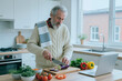 © Seventyfour - Senior Caucasian man preparing healthy meal, slicing eggplant beside laptop in home kitchen. Concept showing active aging, online cooking content, nutrition or retirement lifestyle marketing