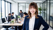© Jesse_4160 - A young professional Japanese woman in business attire smiling at the camera in a modern office with colleagues working in the background