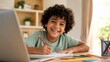 © Space Priest - Happy young boy with curly hair sits at his desk writing in a notebook while using a laptop for schoolwork.