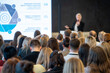 © Anton Gvozdikov - Presenter delivers slide based lecture to diverse audience, professionals listening attentively in conference room setting.