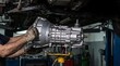 © maxici - Professional mechanic's hands installing a shiny silver car transmission in an auto repair shop. Focus on automotive service, vehicle maintenance, and gearbox repair.