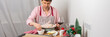 © pundapanda - A man is preparing for a pre-Christmas dinner by baking chicken and potatoes. He is focused on his task while surrounded by festive decorations and a glass of wine, banner