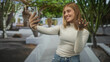 © Krakenimages.com - Woman holds smartphone and waves hand in a white courtyard, smiling for a selfie in casual jeans and turtleneck; happy connection.