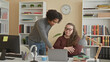 © Krakenimages.com - Man points to laptop and shows papers while seated woman watches with folded arms in an office workspace with bookshelves and a clock; mentoring focus.