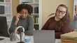 © Krakenimages.com - Man on phone and woman gesturing by laptop at a cluttered desk with headphones and folders in a building; frustration teamwork.