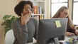© Krakenimages.com - Man at computer yawning with hand covering mouth, seated at desk with monitor and sticky note while coworker types at laptop in a building office with bookshelf and plant; fatigue.