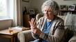 © An - An elderly woman sits in a comfortable armchair, holding her hand to her wrist in a gesture of pain. She wears a cardigan and glasses, and a cup of tea sits on a small table beside her.