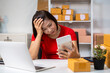 © M+Isolation+Photo - Stressed woman working at a desk with laptop and tablet in a small online shop, surrounded by shipping boxes and packaging supplies.