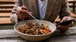 © andyaziz6 - Person holding fork and smartphone while eating a colorful salad from a ceramic bowl at a wooden table
