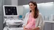 © Liliya - Smiling pregnant woman sitting in a modern clinic, gently holding her belly during a prenatal ultrasound checkup, representing maternity care and health.