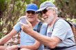 © luciano - Two smiling senior adult men. Wearing caps and backpacks. Are sitting outdoors on a forest trail. Happily looking at a smartphone screen