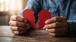 © Eggie - A person's hands hold two halves of a broken red paper heart on a wooden table, symbolizing heartbreak, loss, or the end of a relationship.