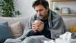 © Jafree - Man covered with blanket sitting on sofa holding steaming cup of tea. Sick person having flu symptoms with cold and fever at home. Healthcare concept for viral illness treatment