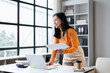 © David - Asian businesswoman smiling, standing at her office desk, holding paperwork, analyzing reports, and using a laptop, embodying professionalism, work life balance, and career success