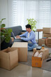 © Pichsakul - Young Asian man working on laptop computer surrounded by cardboard boxes while settling into new apartment with copy space
