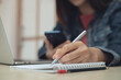 © tippapatt - Business woman working on laptop and smartphone, writing in notebook, business planning, remote work at home, online studying or research