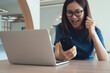 © tippapatt - Excited cheerful young asian woman in casual dress sitting at table, using smartphone and laptop computer, raising hand up and celebrating success at home, close up