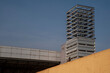 © massimo_g - Un piccione è appollaiato su un muro giallo di fronte a un alto edificio moderno. La torre presenta una struttura geometrica in acciaio con linee orizzontali, in contrasto con il cielo azzurro