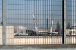 © massimo_g - Recinzione metallica con motivo a griglia in primo piano, che offre la vista di una struttura al di là. La struttura ricorda un ponte sospeso o un elemento architettonico moderno.