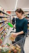 © Lari - Woman uses smartphone to compare prices while grocery shopping with a full cart, planning budget