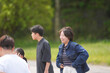© Masakazu Tokashiki - Young woman laughs joyfully outdoors with friends. Group gathers on a path surrounded by greenery. Light, natural setting suggests casual, pleasant gathering. Emotion conveys happiness, connection